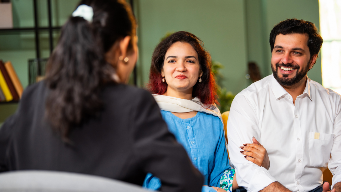 Two people smiling and listening to a woman in a meeting, seated indoors.