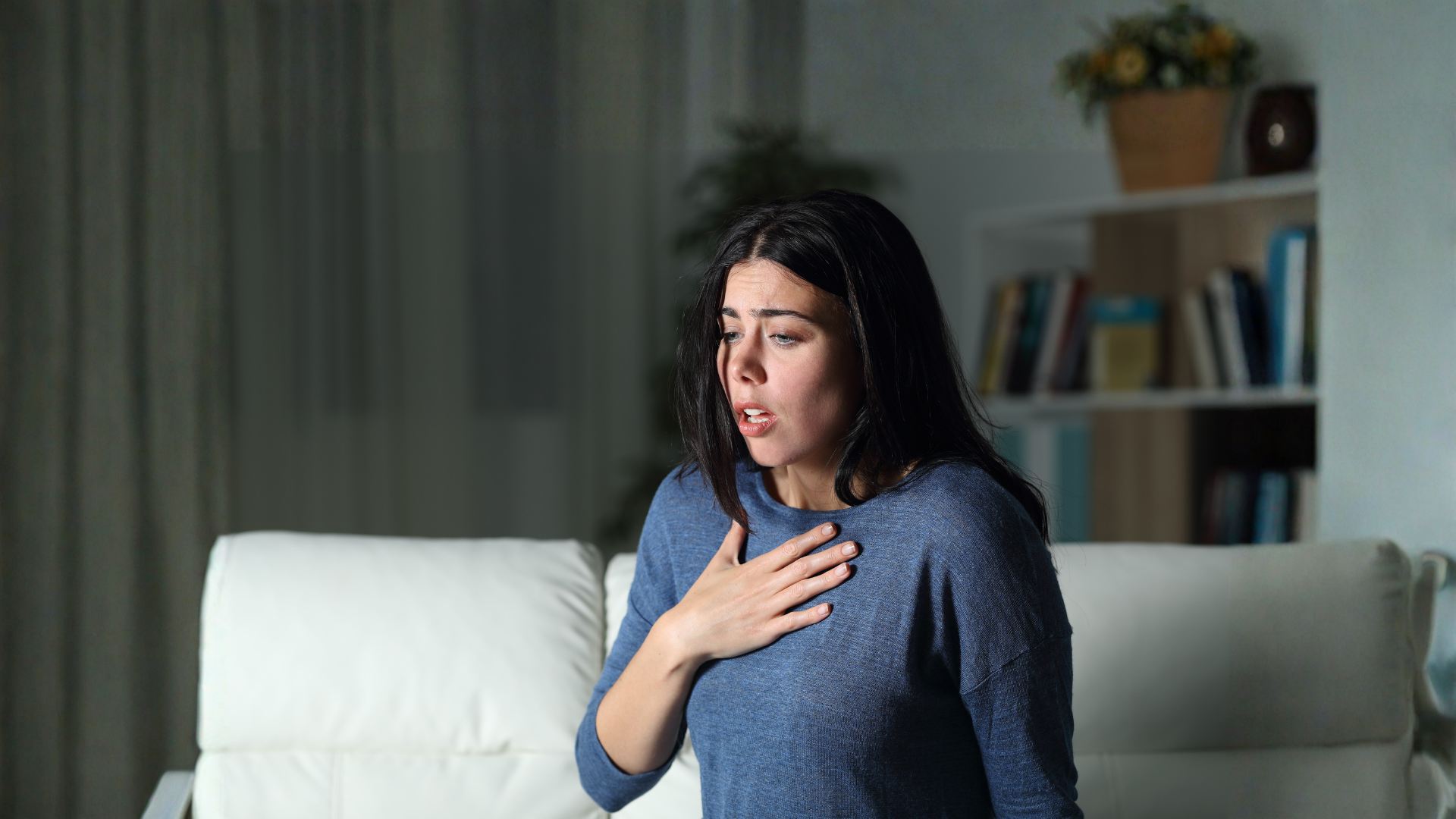 Woman in a blue sweater clutching her chest, looking distressed on a couch.