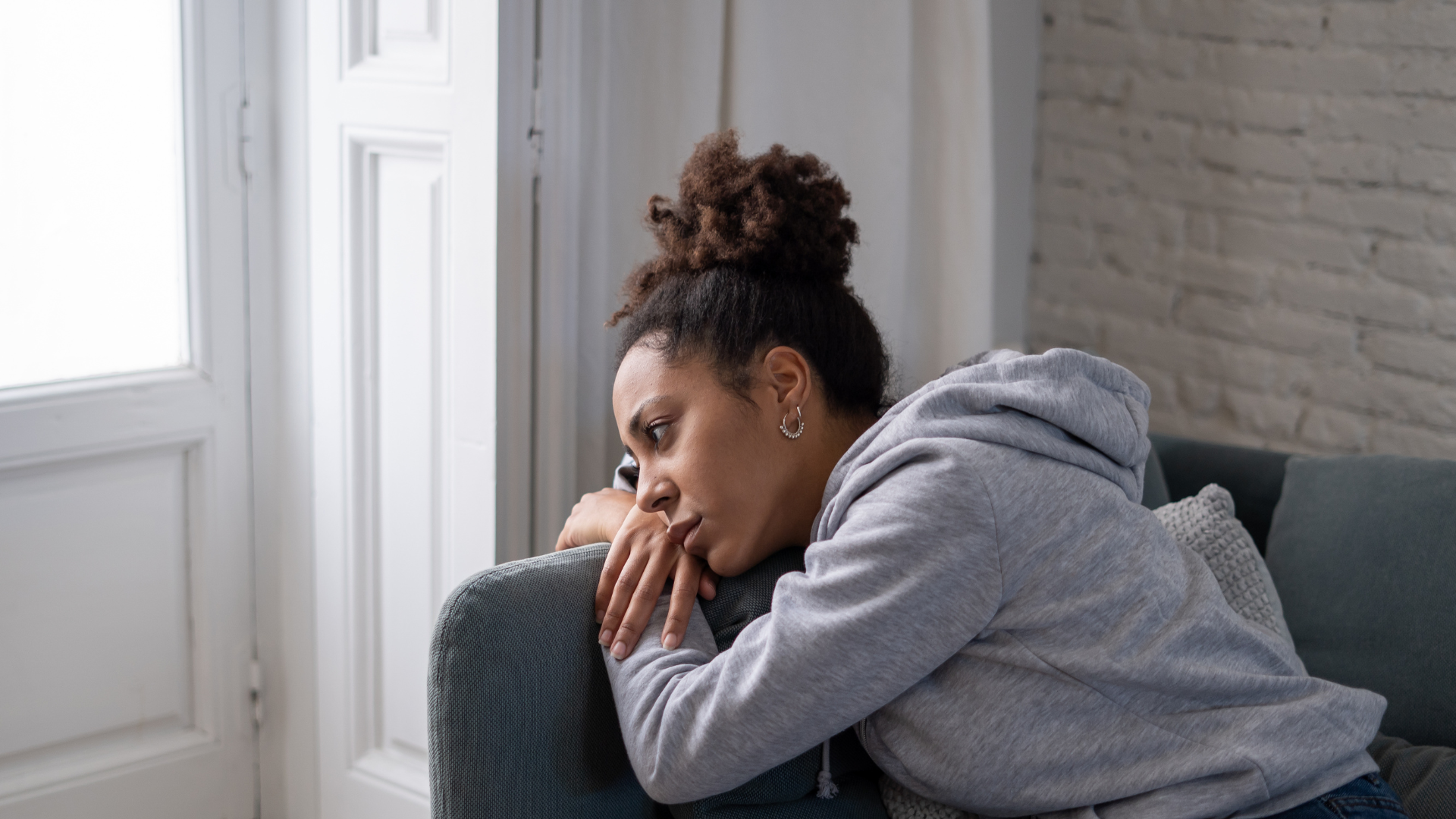 Person in a gray hoodie resting on a couch, looking thoughtful by a bright window