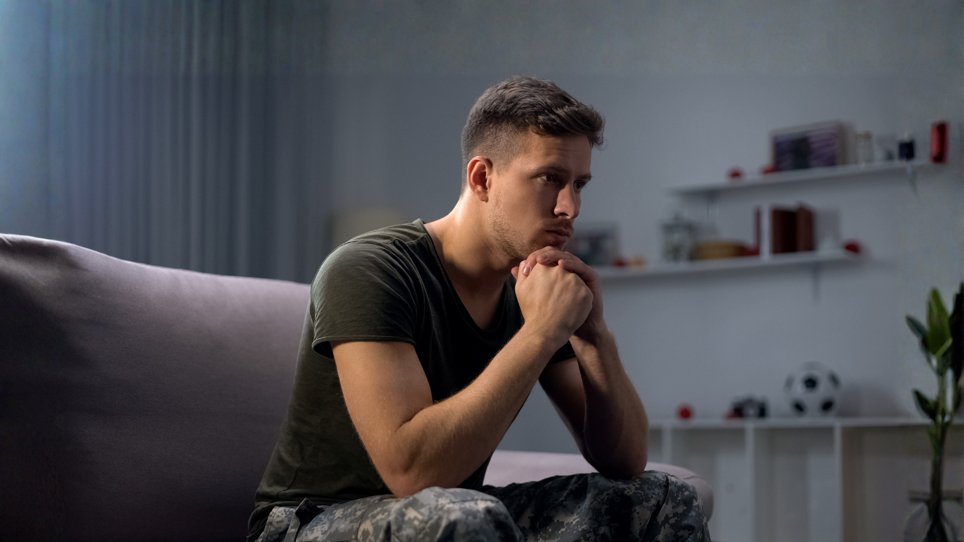 A pensive man sits on a couch, hands clasped, in a softly lit living room.