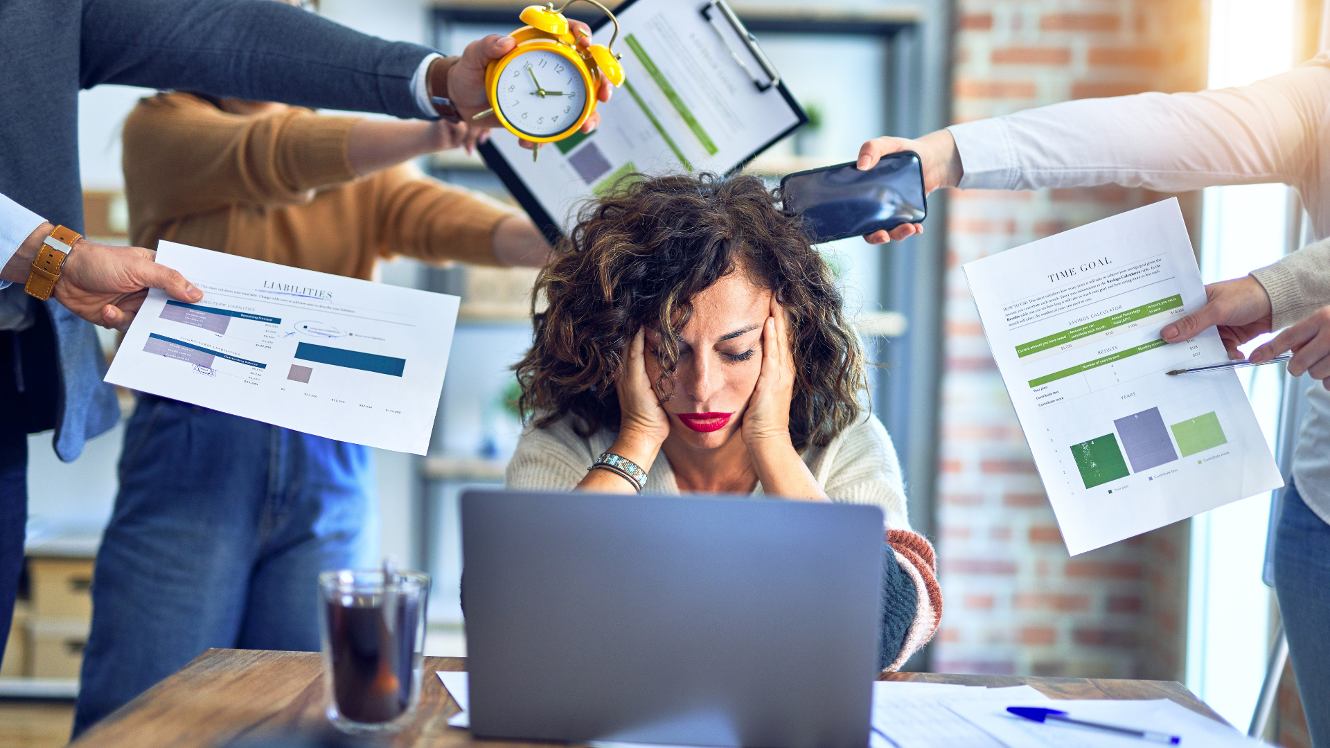 Stressed worker at a laptop while colleagues hold charts and a clock over their desk