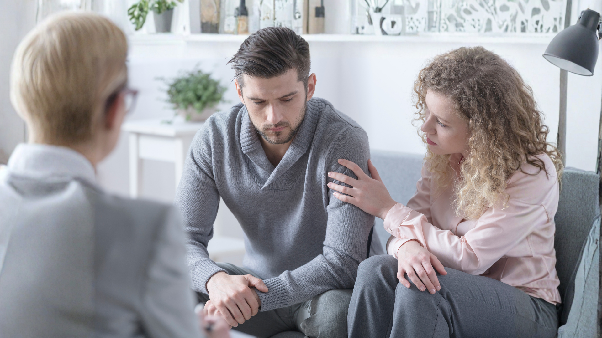 Two people sit close together in a bright room, one comforting the other with a hand on his shoulder.