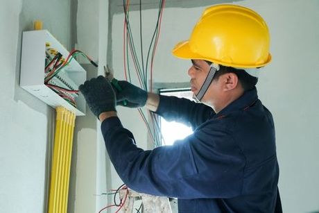 Electrician wiring a wall-mounted junction box, wearing a yellow hard hat and gloves.