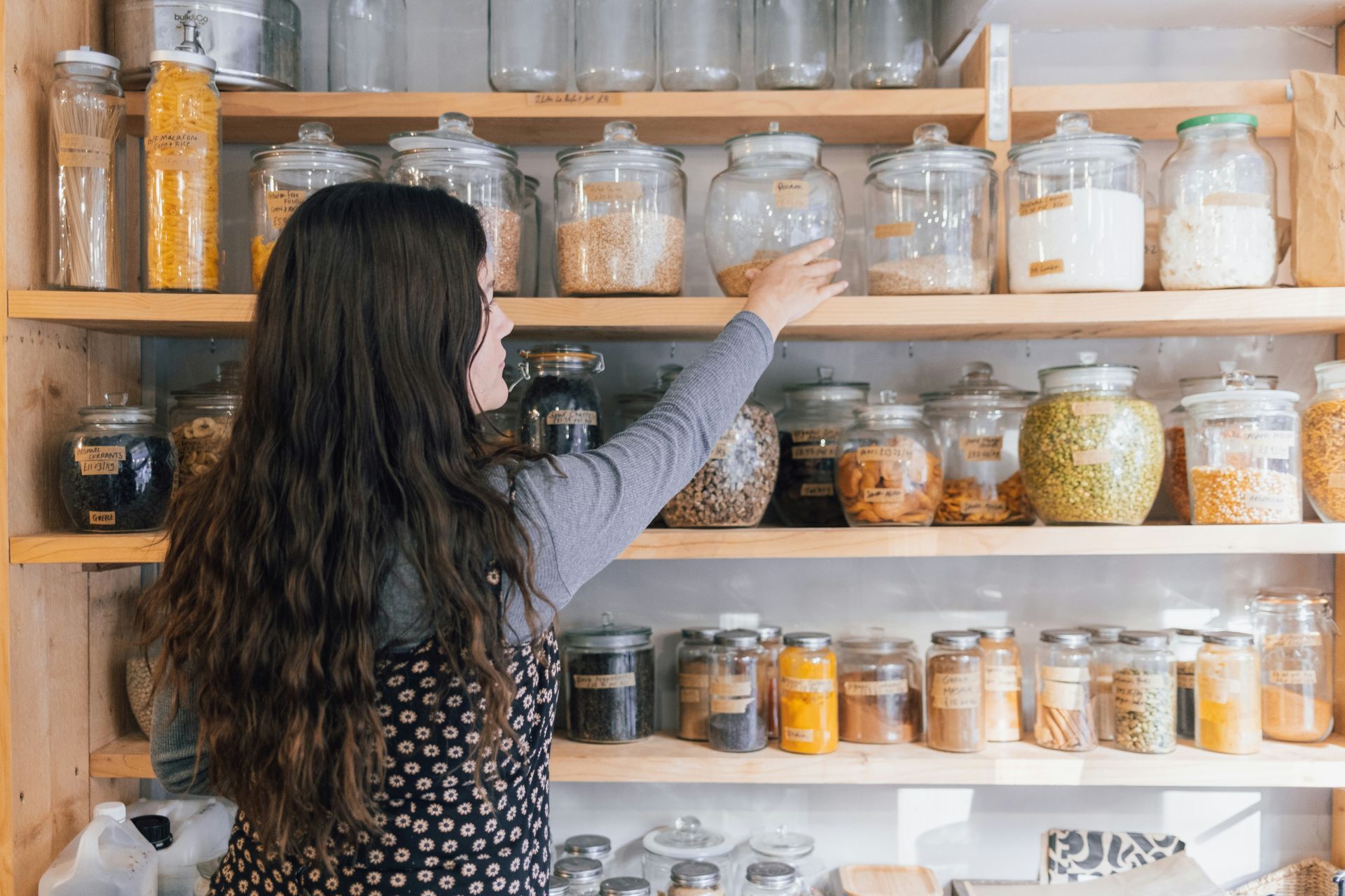 Woman reaching for jars on pantry shelves in a kitchen with rows of glass containers