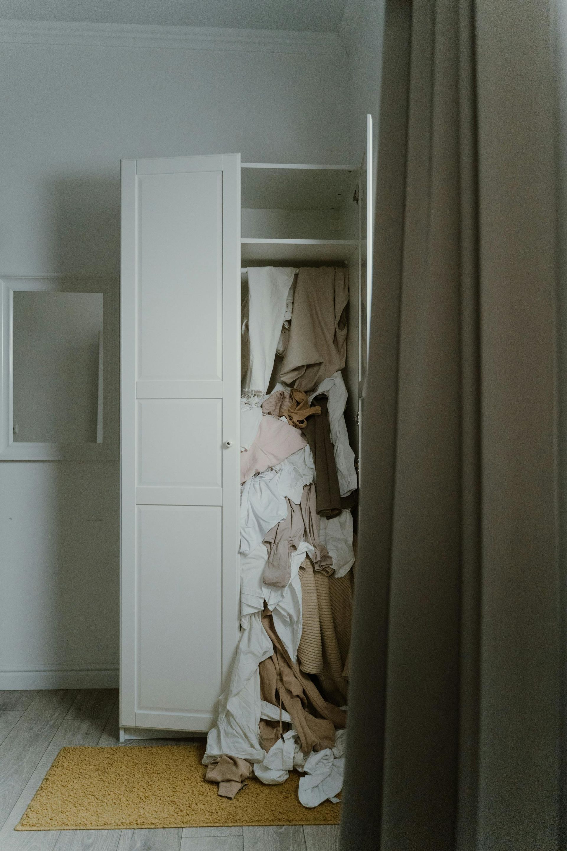 White closet overflowing with clothes; beige curtain at right, yellow rug below.