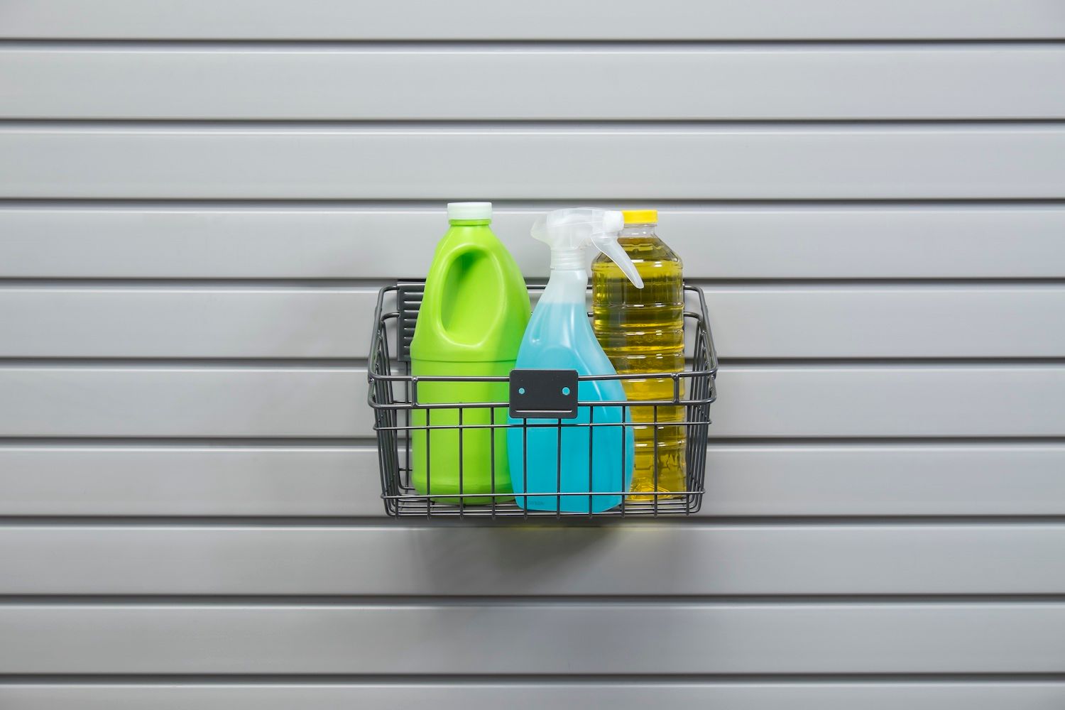 Cleaning supplies in a wire basket mounted on a slatted wall.