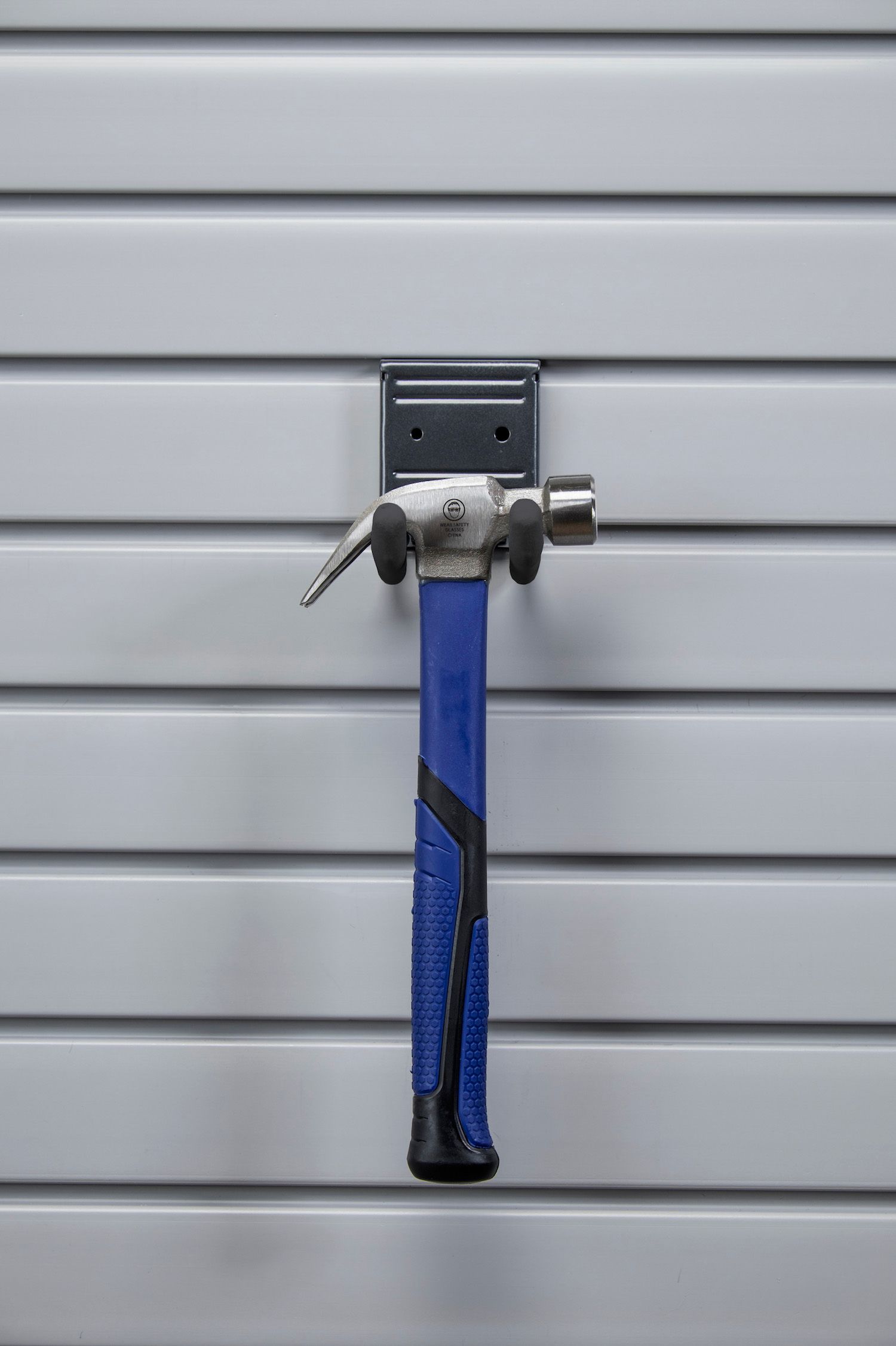 Hammer with blue handle hanging on a gray slat wall.