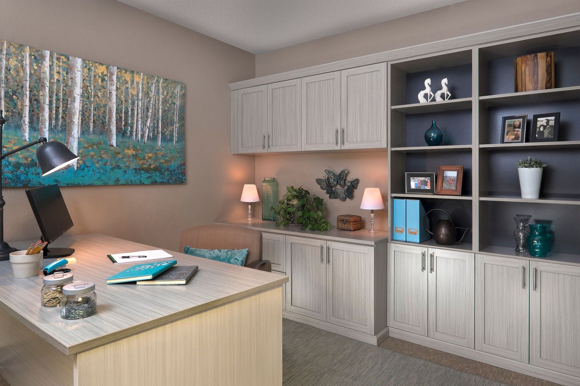 Home office with desk, cabinets, shelves, artwork, and a lamp. Neutral colors and a focus on organization.
