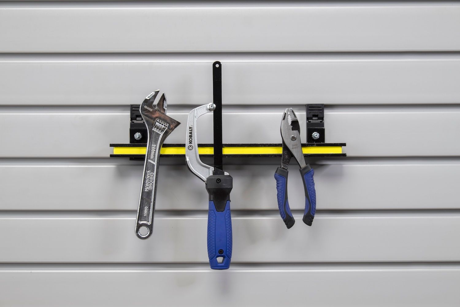 Tools hanging on a gray slatwall: wrench, screwdriver, and pliers on a yellow-accented rail.