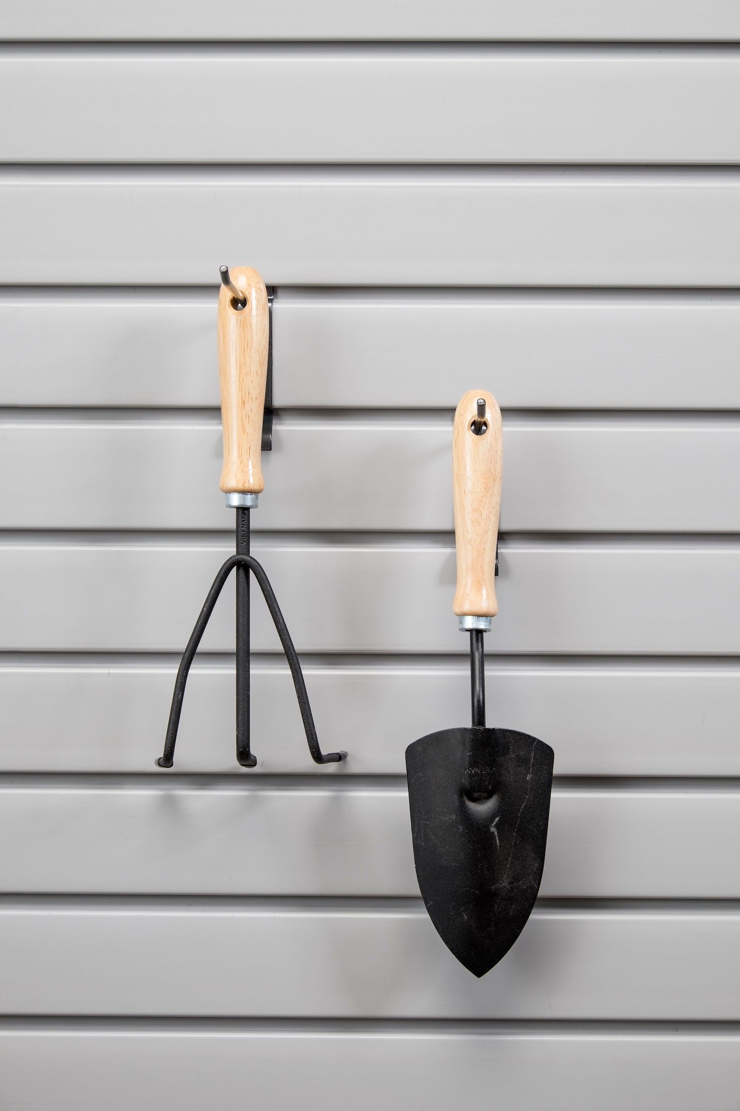 Small garden trowel and hand rake hanging on a gray slatted wall.