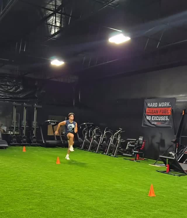 Athlete sprinting through orange cones in an indoor gym with turf and exercise bikes.