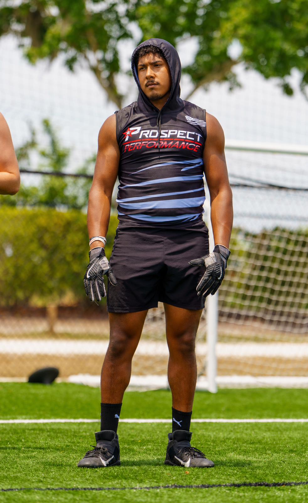 Athlete standing on a soccer field in black training gear and gloves, with a hood up.