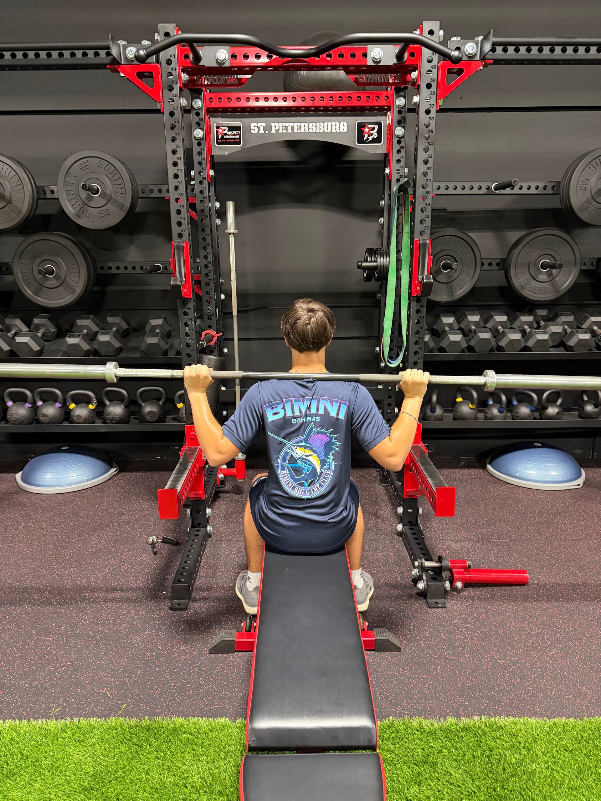 Person seated on a gym machine, gripping handles, facing a large weight rack in a fitness room.