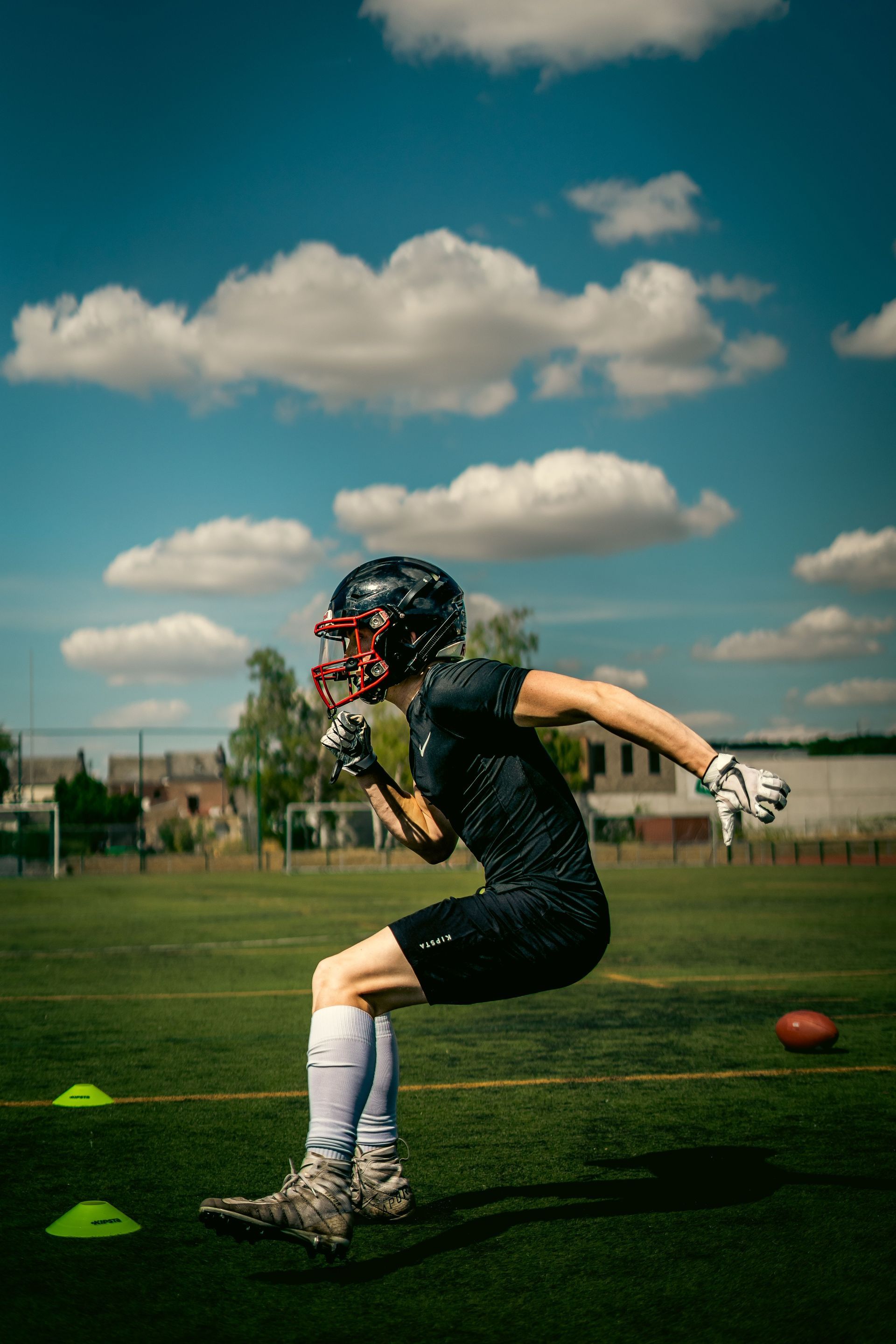 Football player in black running with a ball on a grassy field under a blue sky with clouds