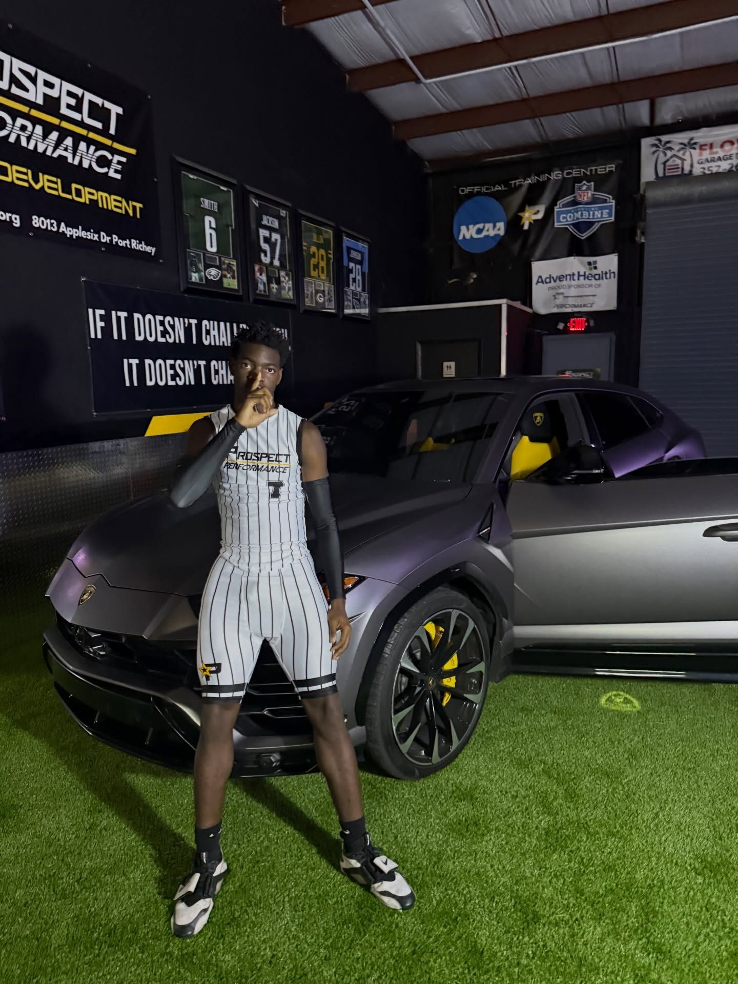 Person in white and black athletic outfit standing in front of a gray sports car on indoor turf.