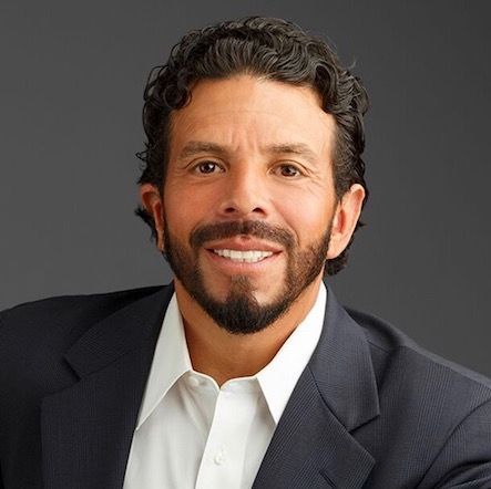 Man in a dark suit, white shirt, smiling, with dark hair and beard, against a gray background.