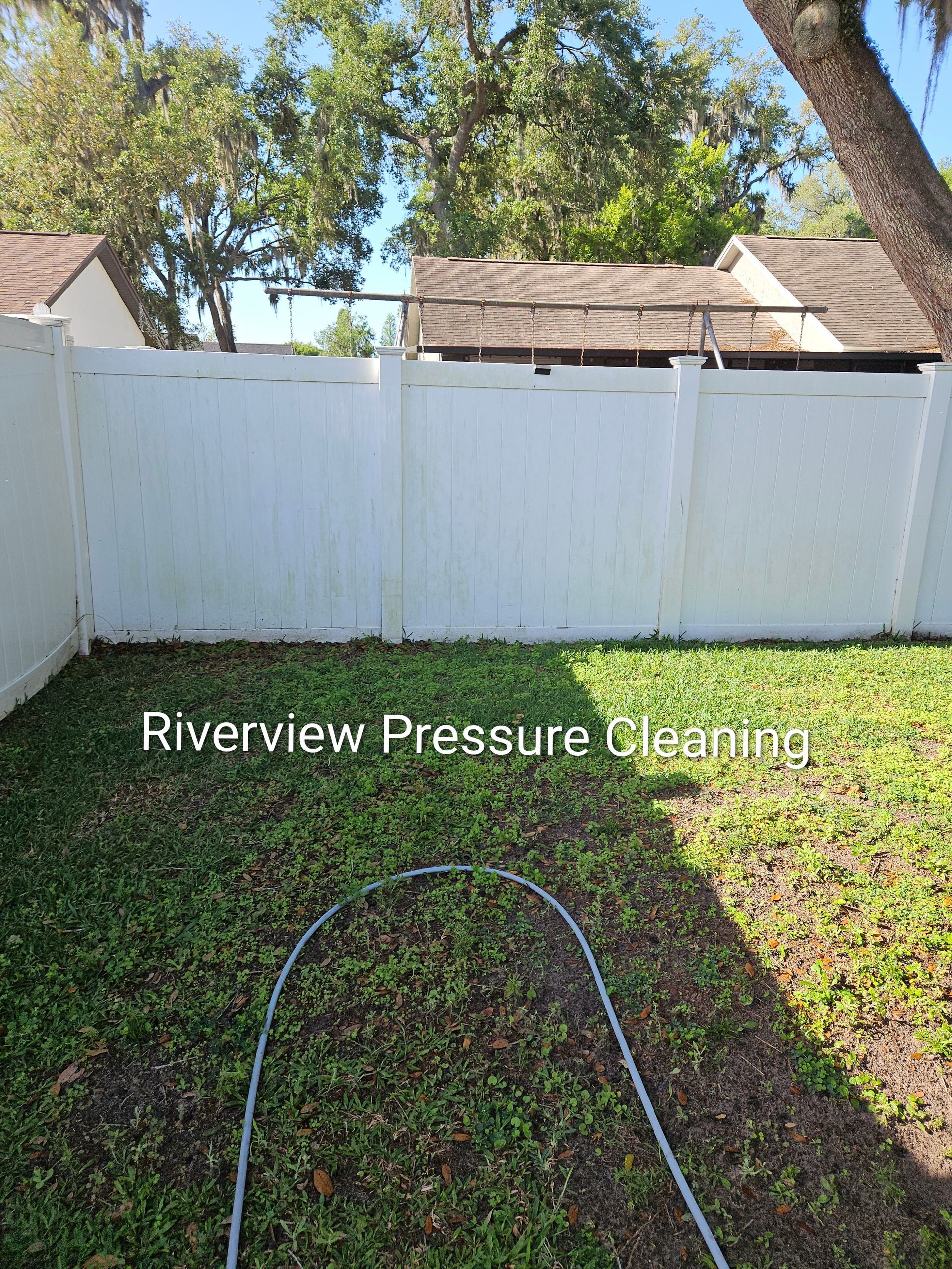 Backyard with white fence, trees, and a pressure washing hose on grass, labeled “Riverview Pressure Cleaning”