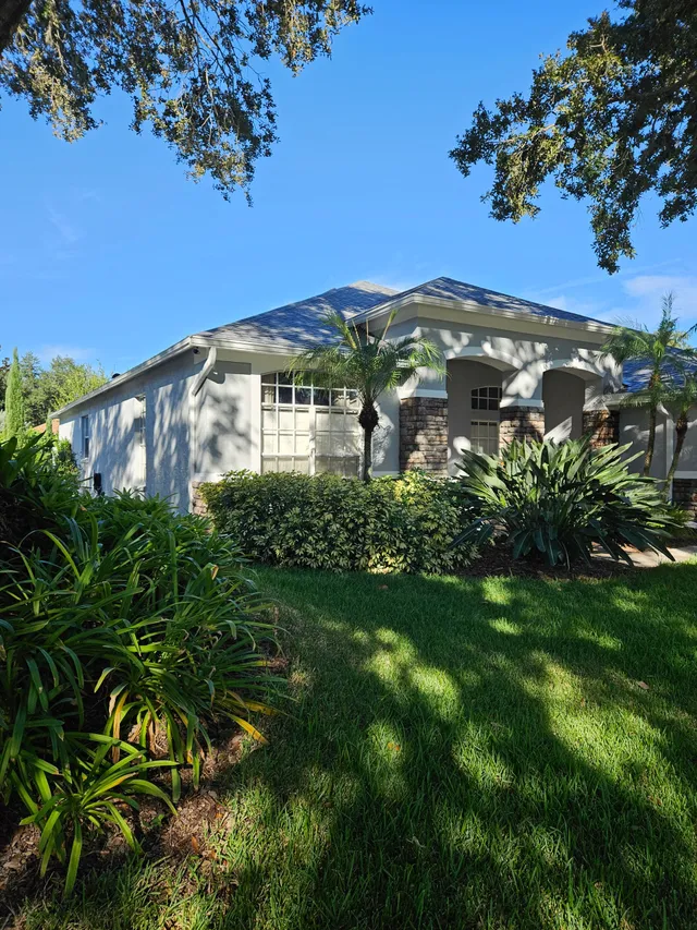 Single-story white house with a front porch, surrounded by lush green trees and shrubs under a blue sky