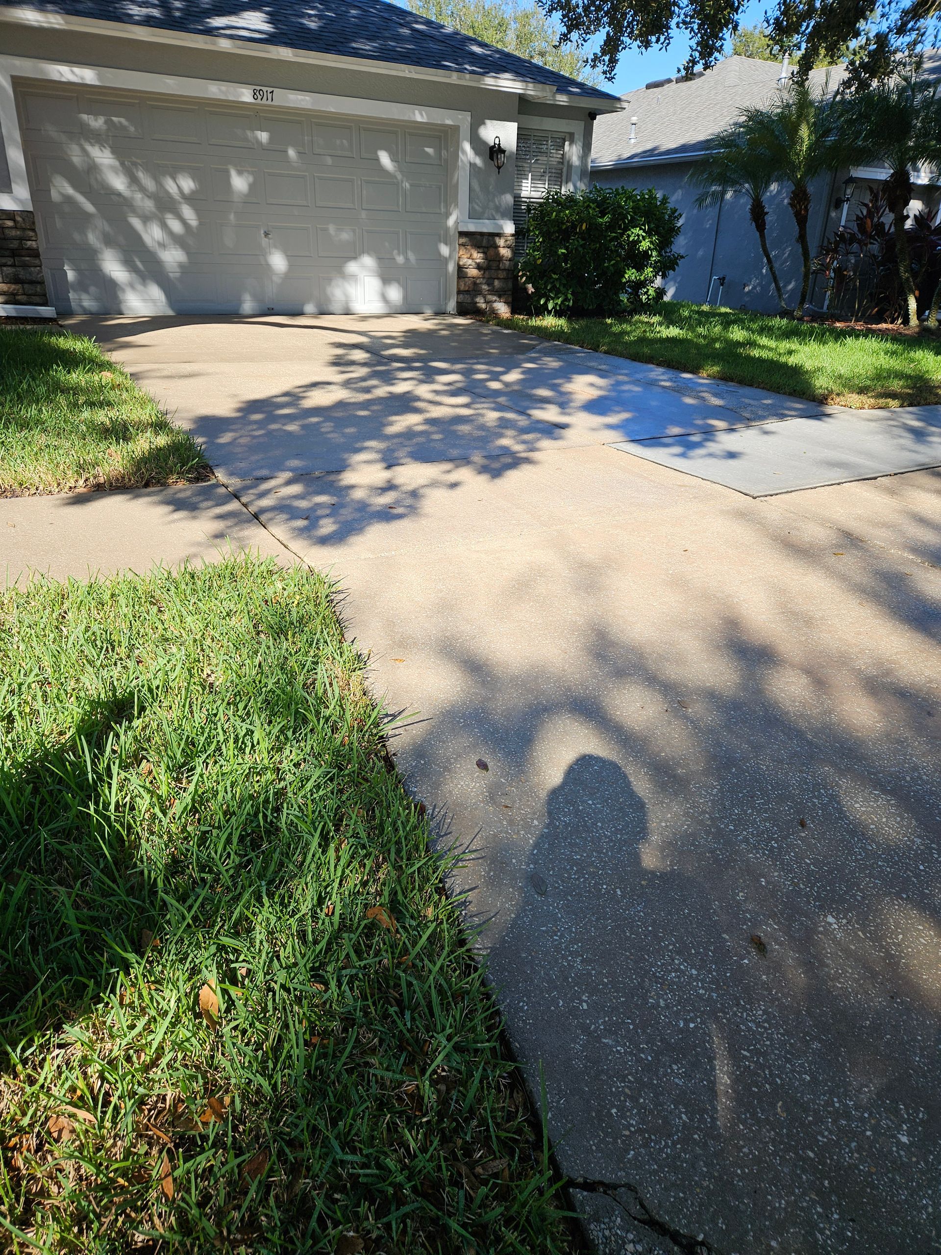 Driveway leading to a beige garage in a sunny residential yard with grass and a blue fence.