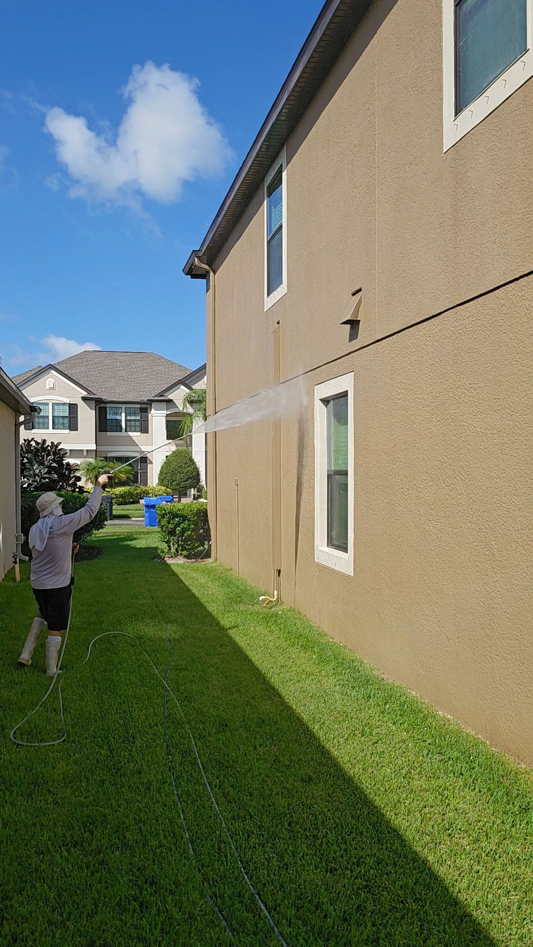 Person in a grassy backyard reaching toward a beige house wall on a sunny day