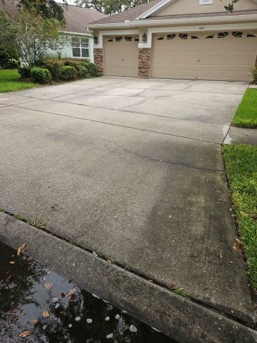 Concrete driveway in front of a two-car garage. Gray concrete with dark streaks and cracks, green lawn on the right.