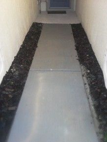 Narrow concrete walkway between white walls, lined with dark mulch on both sides.