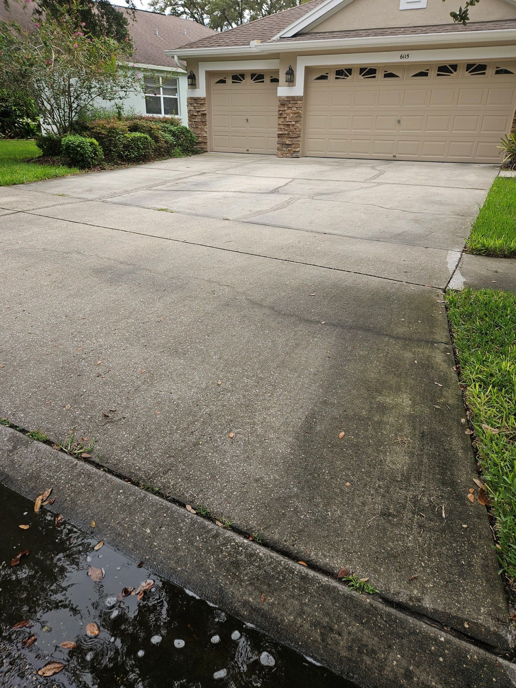 Concrete driveway leading to a beige garage beside a house and front garden, with a wet curb in the foreground