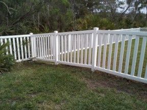 White picket fence with a gate along a grassy yard, bordered by trees and shrubs.