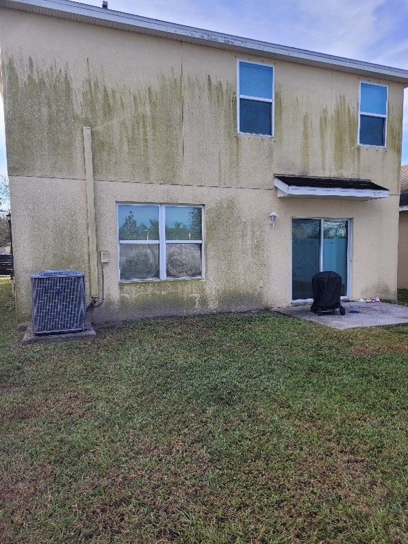 Back of a beige two-story house with stained walls, grassy yard, and a small covered patio.