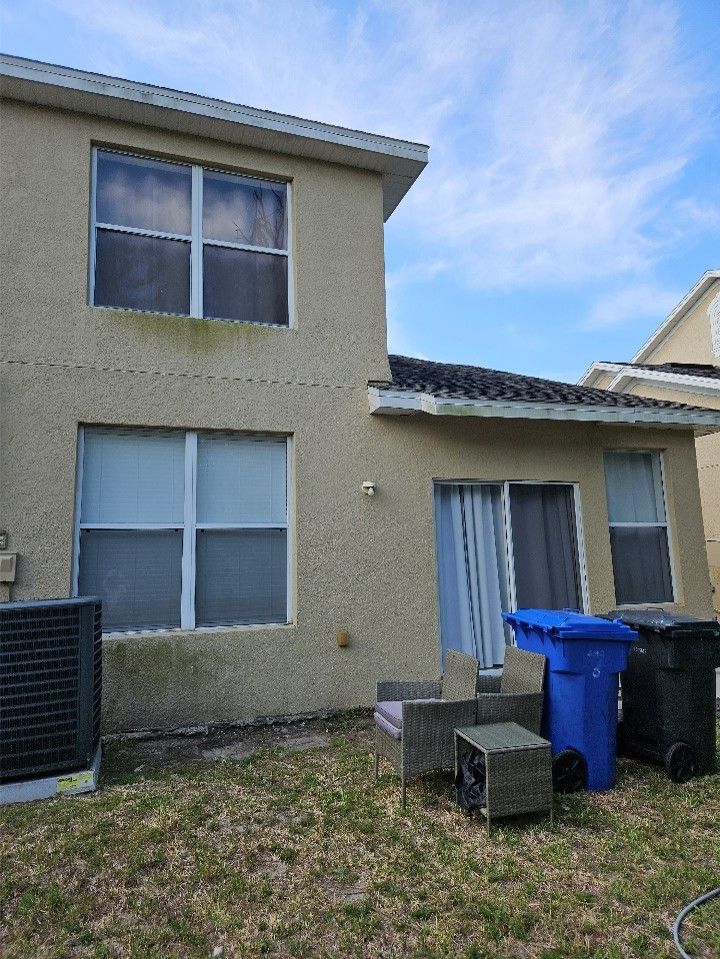 Rear view of a beige two-story house with windows, patio furniture, and blue trash bins on a grassy yard.
