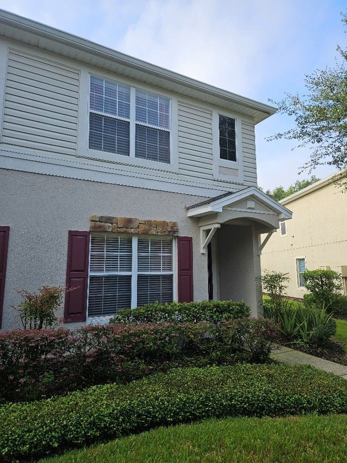 Two-story suburban house with white siding, maroon shutters, and manicured hedges under a blue sky