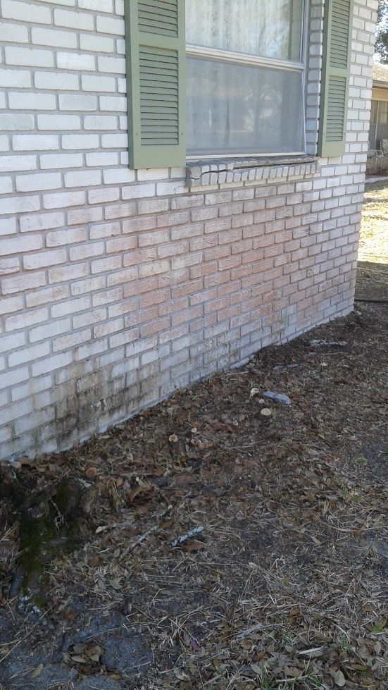 Brick house exterior with a window and green shutters above a bare dirt patch by the foundation