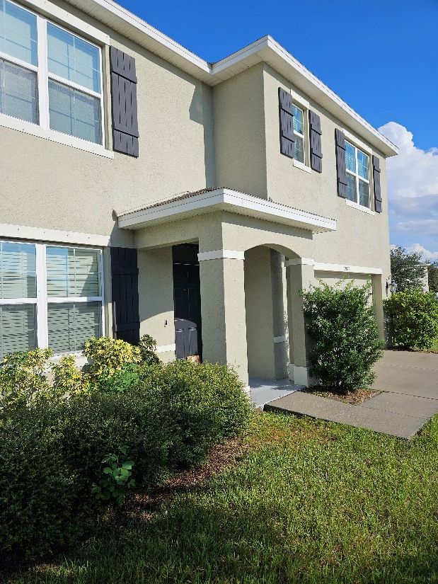 Two-story beige townhouse with a small covered entry, shrubs, and a front lawn in bright sunlight