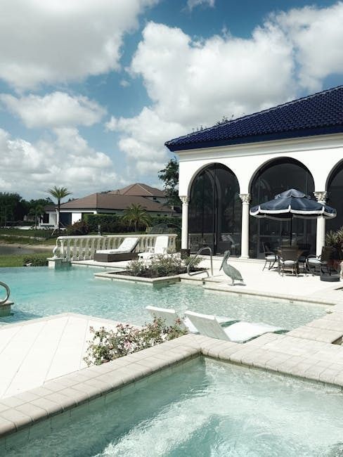 Luxury pool area with white architecture, blue roof, pool, and spa under a cloudy sky.