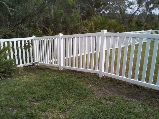 White picket fence bordering green grass, with trees in the background.
