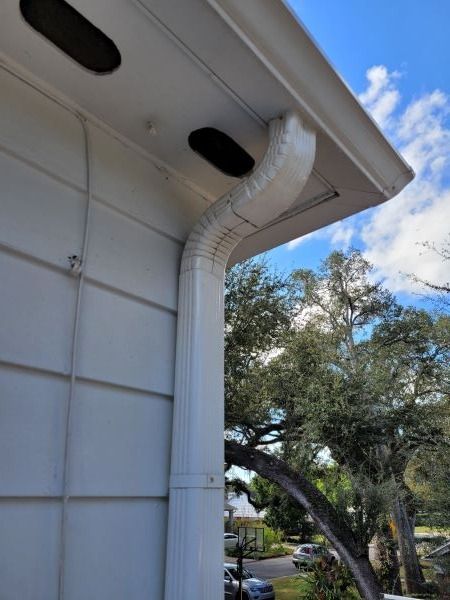 White rain gutter extending from a house's roof. Clear sky and trees are in the background.