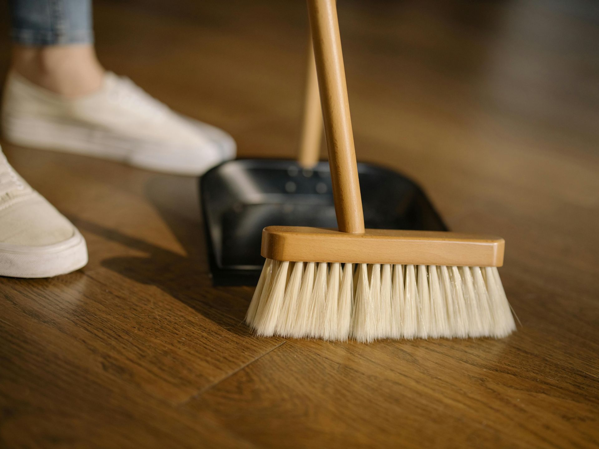 Broom and dustpan on a wooden floor, next to a person's feet in white shoes.