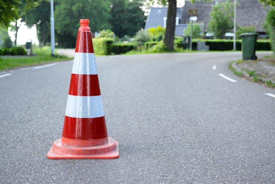 Traffic cone on an asphalt road, painted red and white. Houses and greenery are in the blurred background.