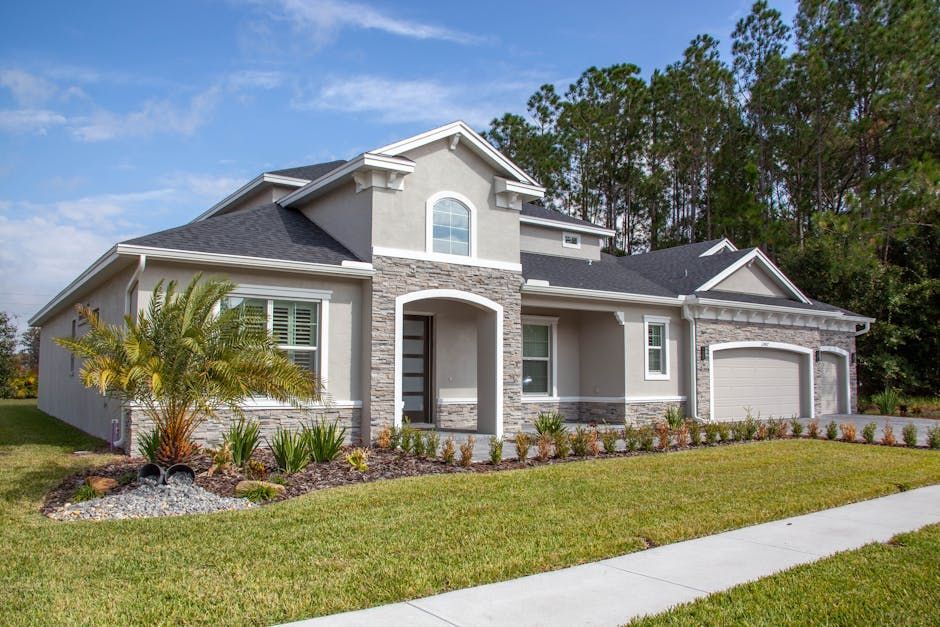 Modern home with light gray stucco, stone accents, and a manicured lawn under a blue sky.