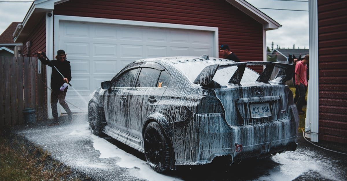 A dark gray car being washed with soap outside a garage; two people are hosing it down.