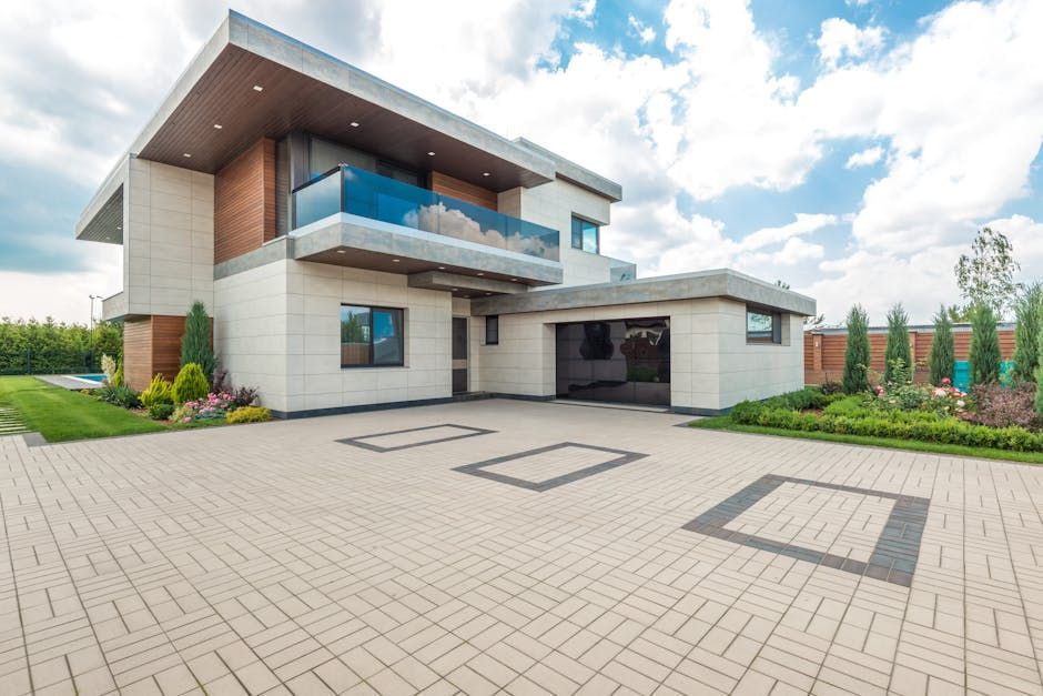 Modern two-story house with a light stone facade, glass balcony, and paved driveway on a sunny day.