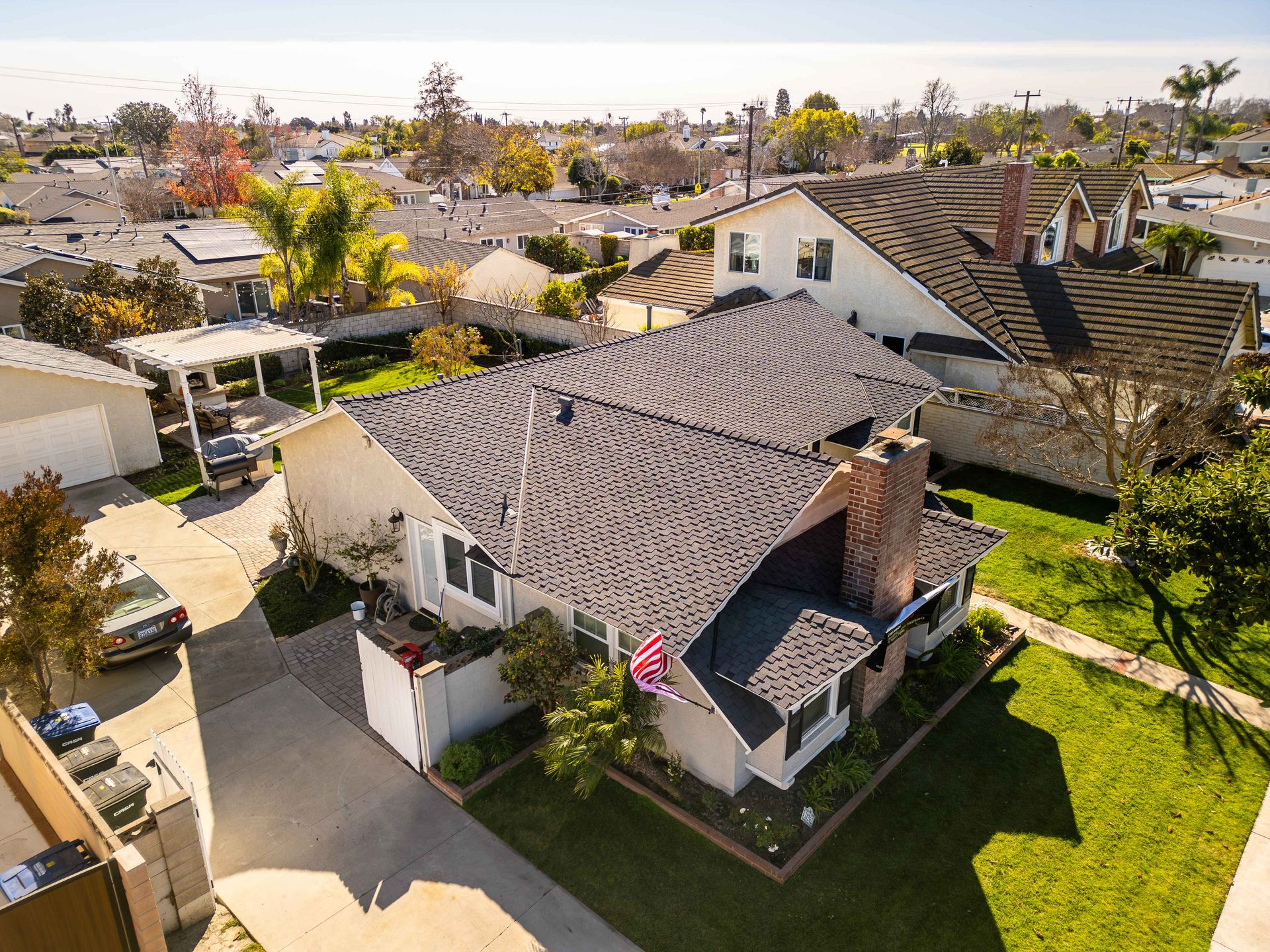 Aerial view of a suburban house with a dark roof and a brick chimney, surrounded by green lawns and other houses.