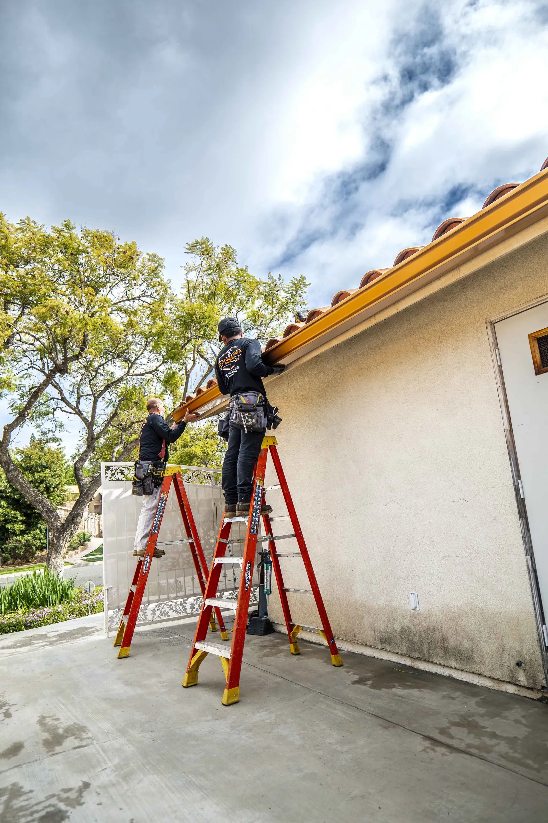 Two workers on ladders install gutters on a white building under a cloudy sky.