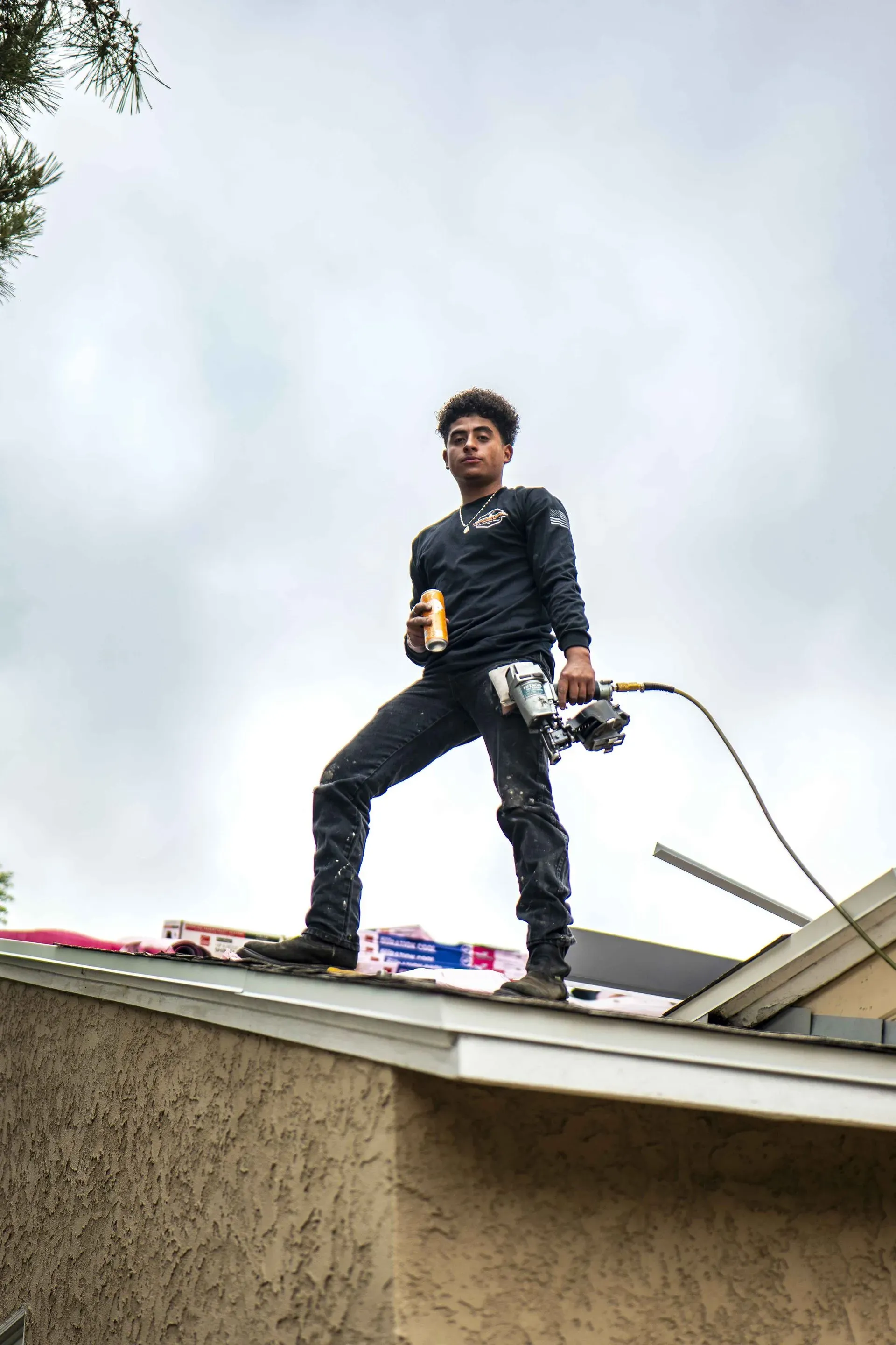 Man on a rooftop holding a can and a nail gun under cloudy sky.
