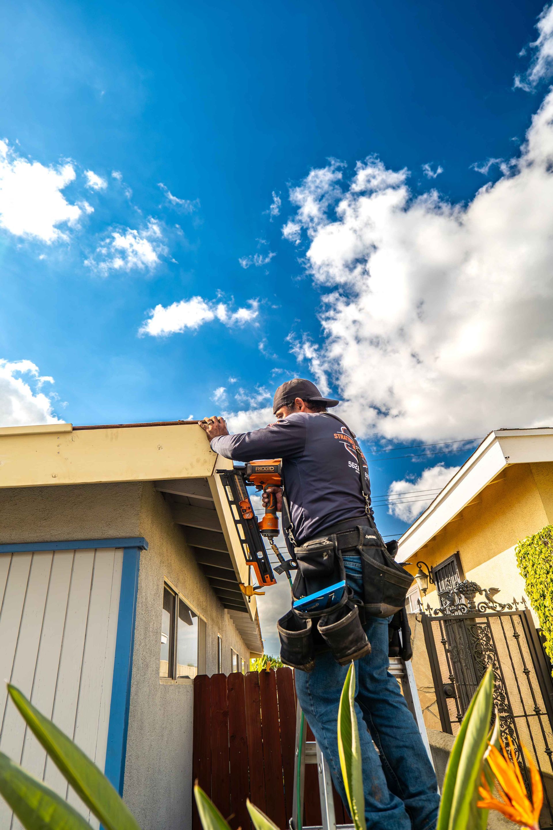 Roofer on a ladder installing shingles on a house roof under a blue sky with fluffy clouds.