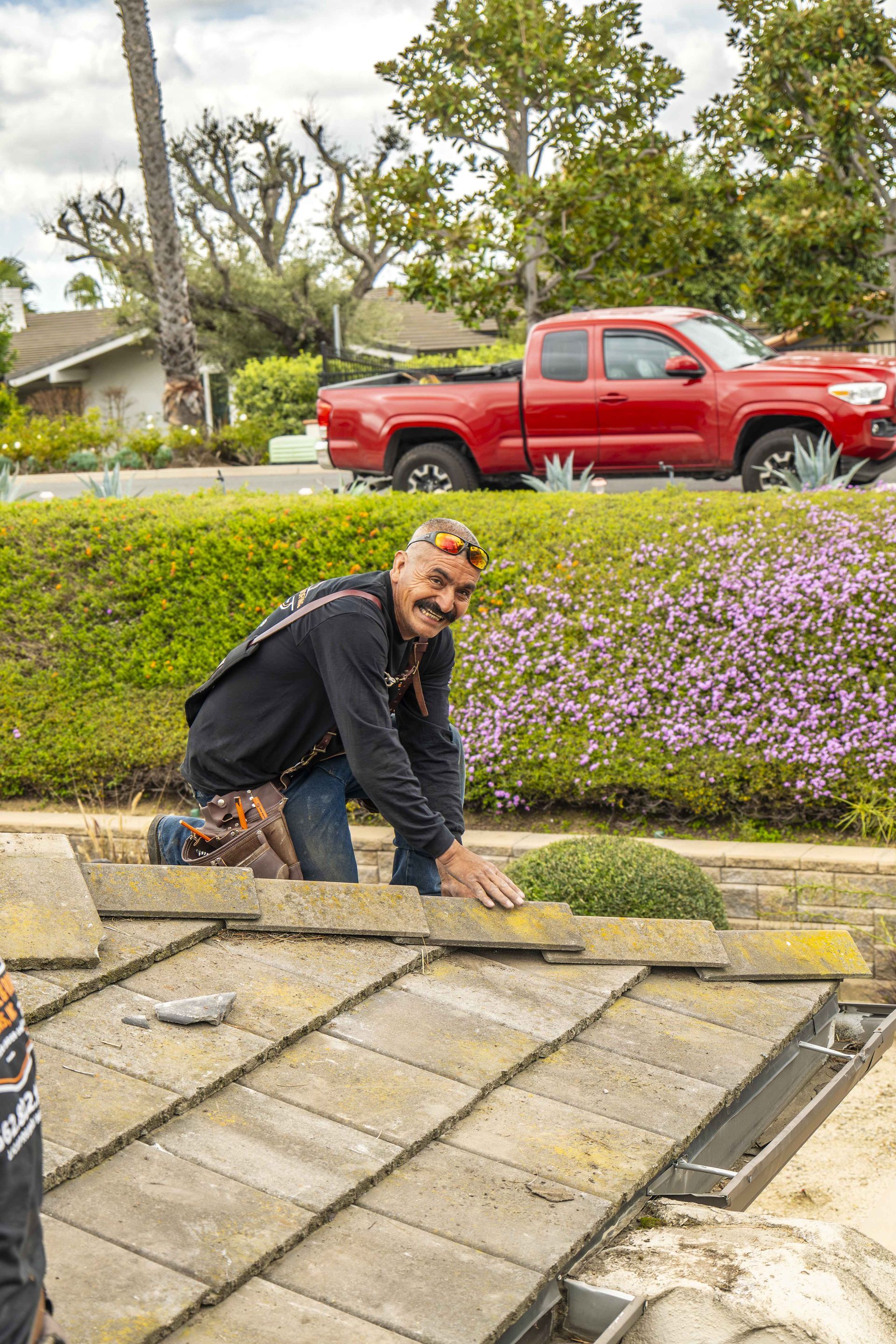 Man on roof with red truck in background. Kneeling, smiling, inspecting tiles. 