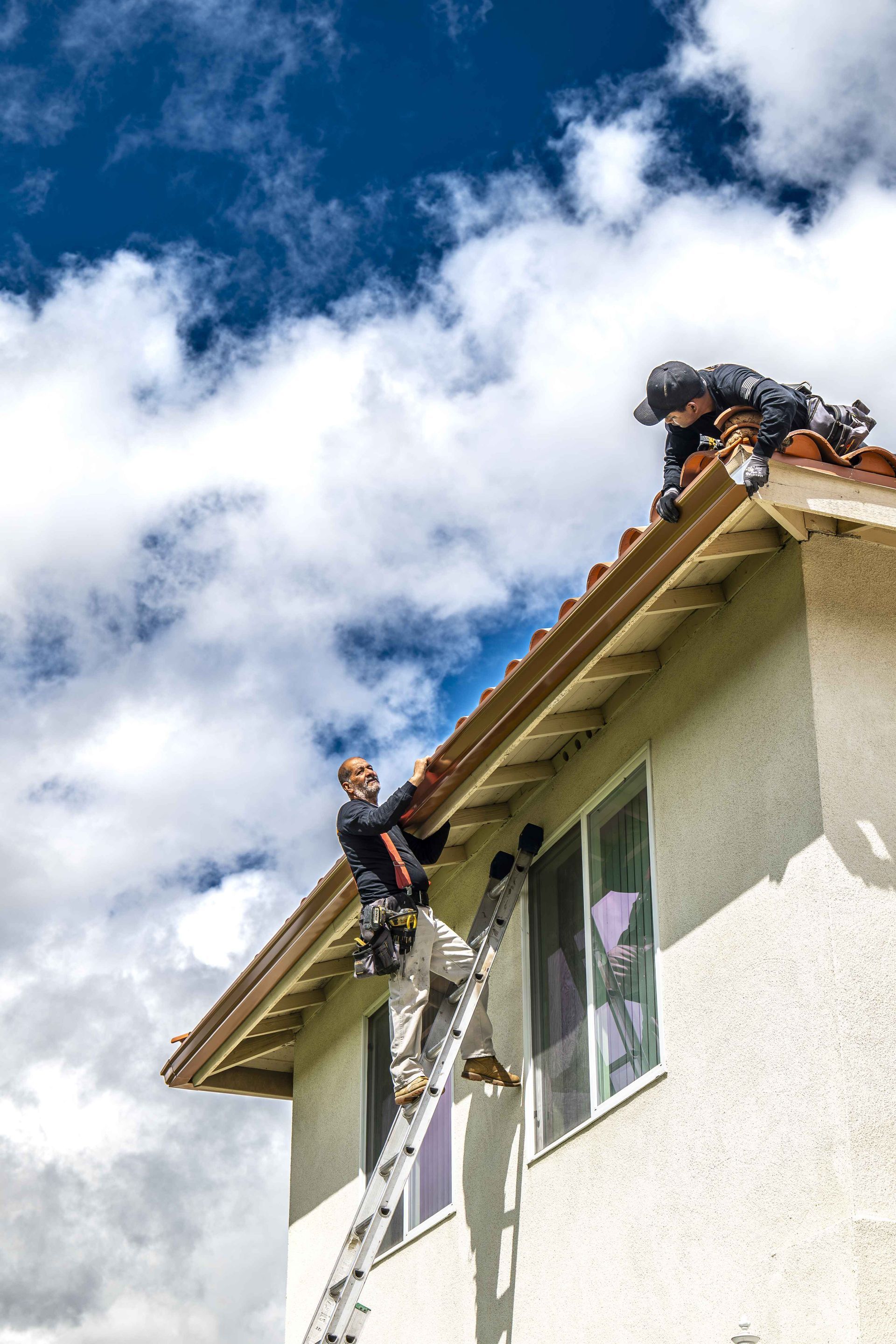 Two workers on a ladder and roof repairing gutters on a house under a cloudy sky.