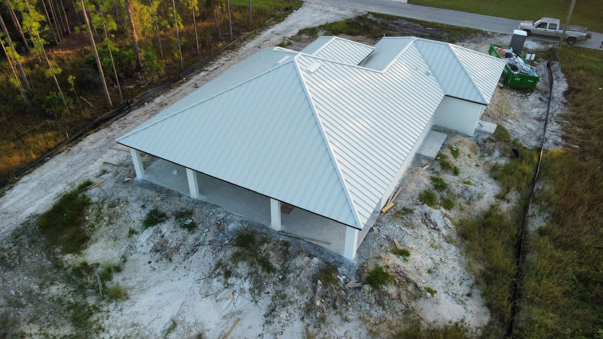 New house under construction with a light-colored metal roof and concrete supports, on a sandy lot.