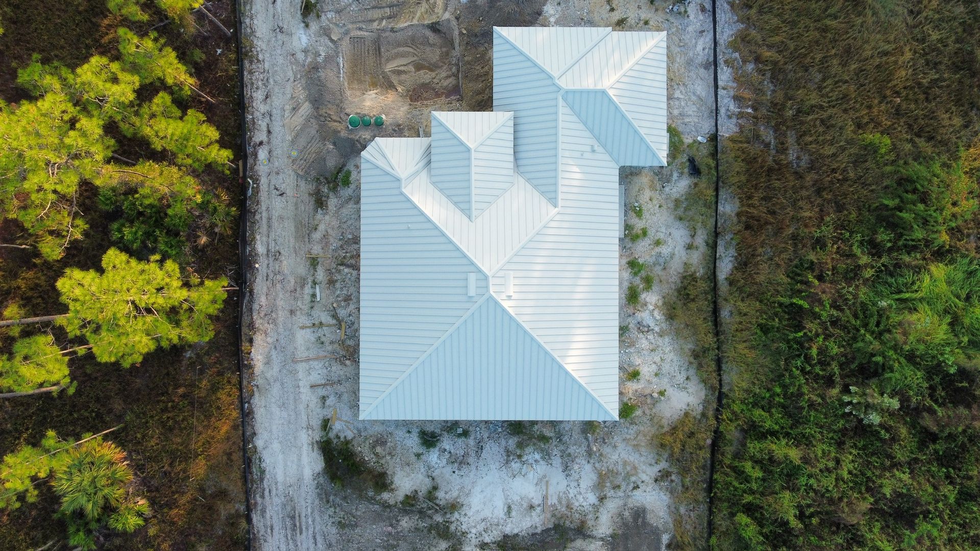 Overhead view of a house with a complex, light-colored roof amidst dirt and trees.