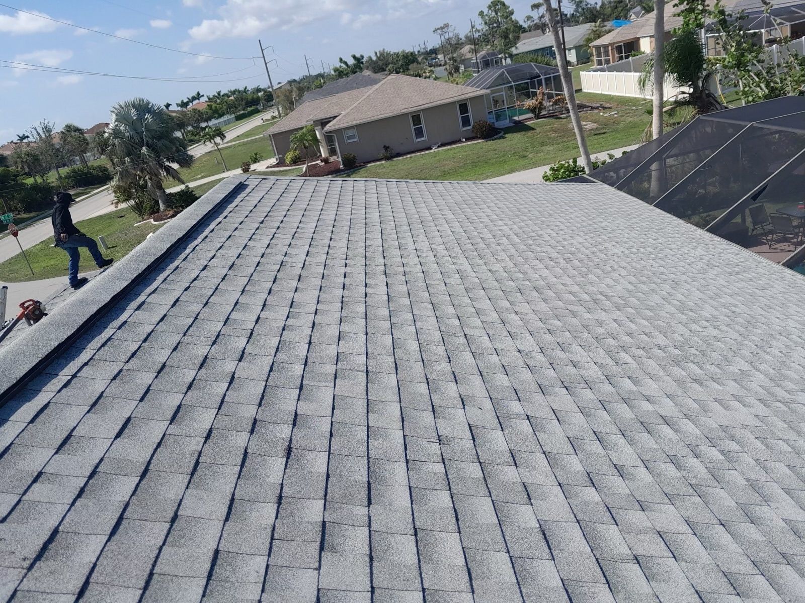 A roof with gray shingles, viewed from above, with a worker on the side in the background.